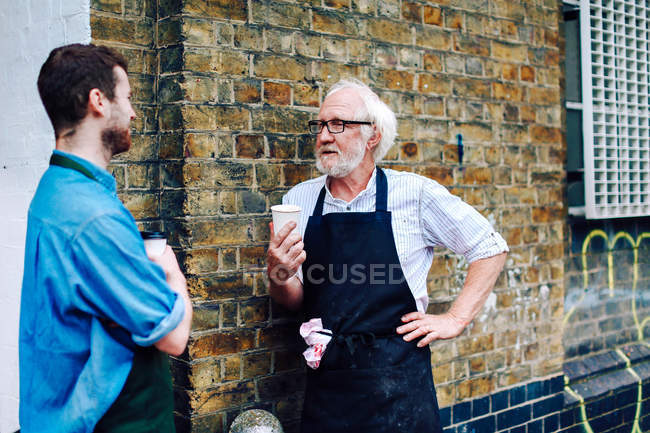 Senior craftsman drinking coffee and chatting to young man outside workshop — Stock Photo