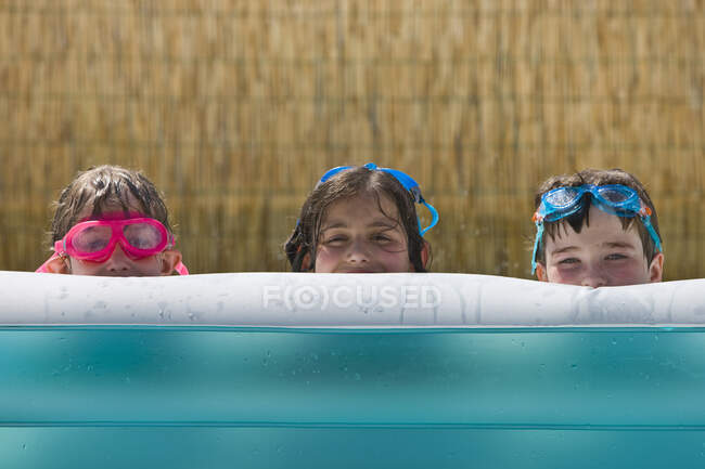 Portrait of two girls and boy peering from garden paddling pool — Stock Photo