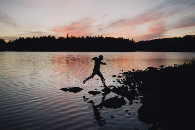 Чоловік стрибає на камені на краю води, сутінки — стокове фото