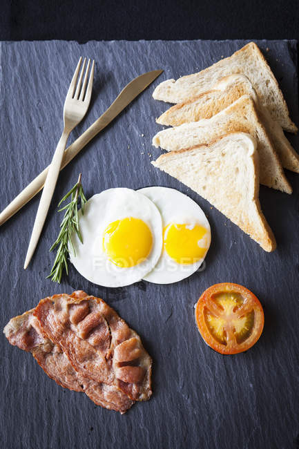 Vista superior de la tostada con tocino, huevos fritos y desayuno de tomate en pizarra - foto de stock