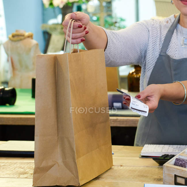 Tiro cortado de assistente de vendas entregando saco de compras e recibo na loja de presentes checkout — Fotografia de Stock