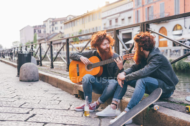 Young male hipster twins with red hair and beards playing guitar on canal waterfront — Stock Photo