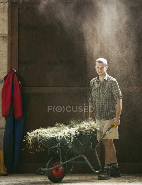 Portrait of young male farmworker with wheelbarrow in dusty farm barn — Stock Photo