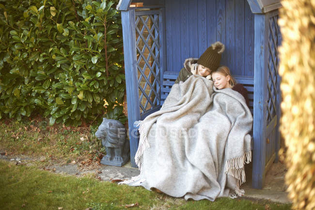 Братья и сёстры, завернутые в одеяло, отдыхают на скамейке — стоковое фото