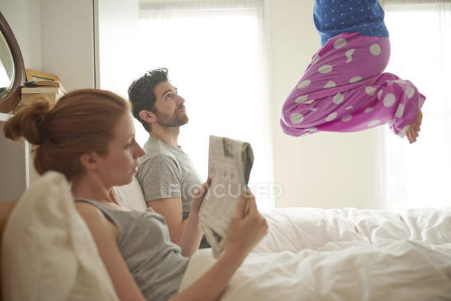 Взрослая пара читает широкополосный лист, пока дочь прыгает на кровать — стоковое фото