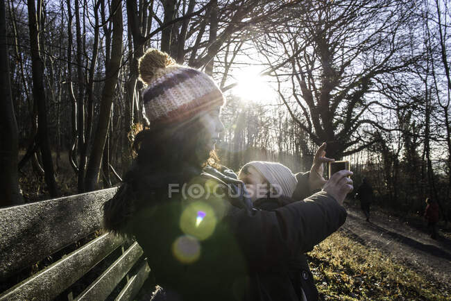 Mother sitting on bench with baby boy, taking selfie with smartphone — Stock Photo