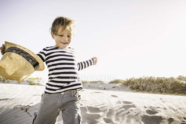 Menino correndo ao longo da praia, segurando chapéu de palha — Fotografia de Stock