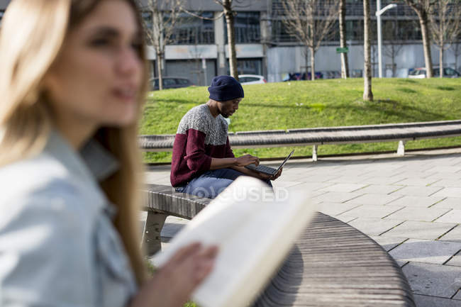 Students reading book and using laptop while sitting on wooden bench in park — Stock Photo