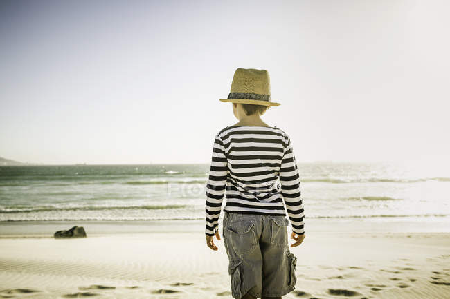 Niño de pie en la playa, mirando al mar, vista trasera - foto de stock