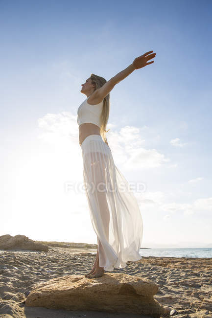 Молода жінка стоїть на скелі на пляжі, руки витягнуті, низький кут зору — стокове фото
