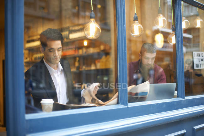 Vista janela de empresários lendo jornal e usando laptop no café — Fotografia de Stock