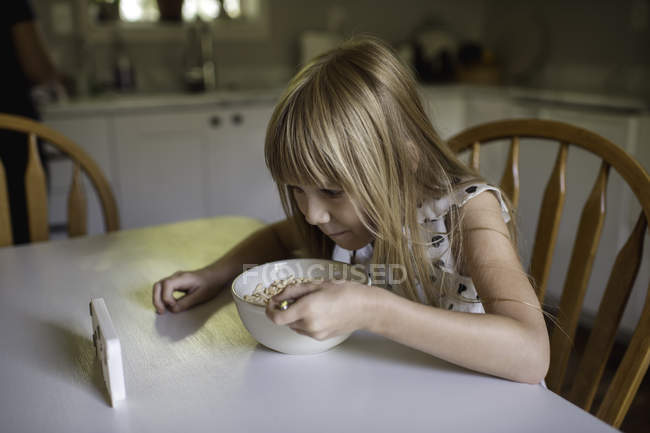 Petite fille manger le petit déjeuner tout en regardant dispositif électronique — Photo de stock