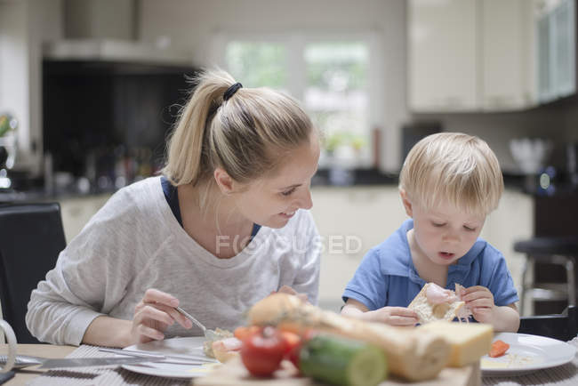 Madre e hijo sentados a la mesa comiendo juntos - foto de stock