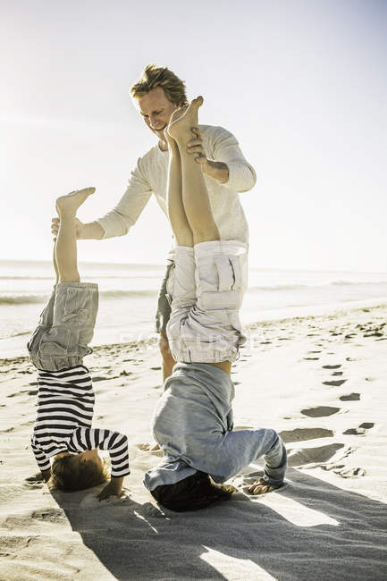 Father helping sons with handstand on beach — Stock Photo