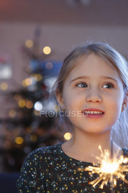 Mädchen vor dem Weihnachtsbaum hält Wunderkerze und schaut lächelnd weg — Stockfoto