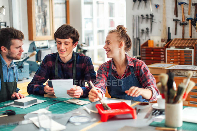 Junge Kunsthandwerkerin hält Pinsel in der Hand und lacht und lächelt mit zwei jungen Kunsthandwerkern im kreativen Druckatelier — Stockfoto