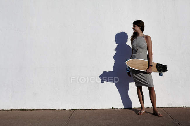 Jovencita guay parada frente a la pared blanca sosteniendo el monopatín, Nueva York, Estados Unidos - foto de stock