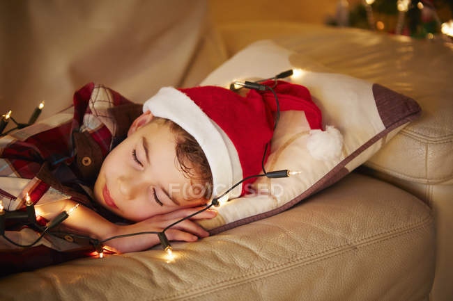 Boy sleeping, wearing santa hat and lights on sofa at christmas — Stock Photo