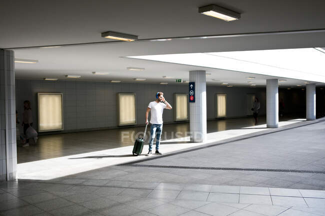 Молодий чоловік на вокзалі, тягне колісну валізу, носить навушники, використовуючи смартфон — стокове фото
