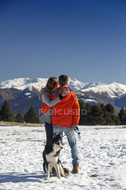 Vista trasera de pareja romántica con perro mirando al paisaje de montaña cubierto de nieve - foto de stock