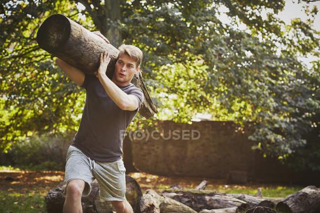 Молодий чоловік тренується, піднімаючи дерев'яний багажник на плечі в парку — стокове фото