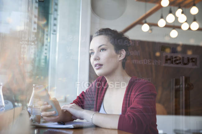 Young woman sitting in cafe, holding smartphone, looking out of window — Stock Photo
