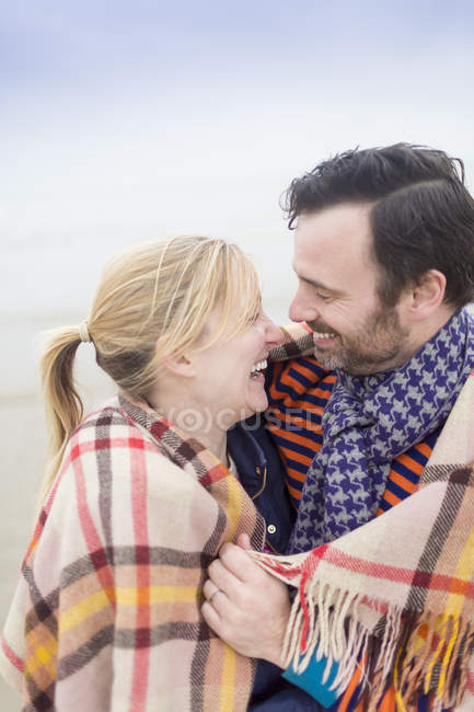 Casal na praia rindo e mantendo-se quente sob cobertor — Fotografia de Stock