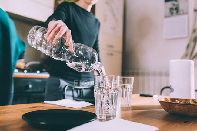 Cortado tiro de mujer joven vertiendo agua en la mesa de la cocina - foto de stock
