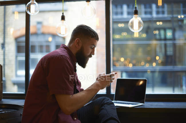 Young male hipster looking at smartphone in cafe window seat — Stock Photo