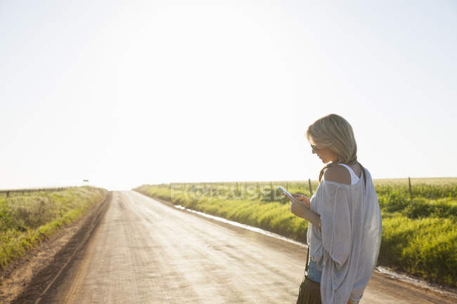 Mulher adulta média andando na estrada do país, olhando para o telefone móvel — Fotografia de Stock