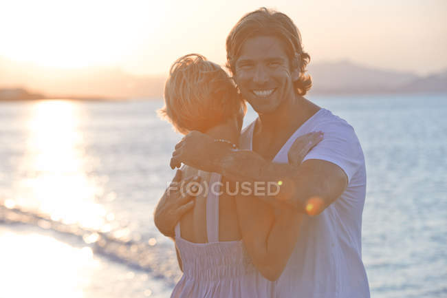 Couple hugging on beach at sunset, man looking at camera and smiling — Stock Photo