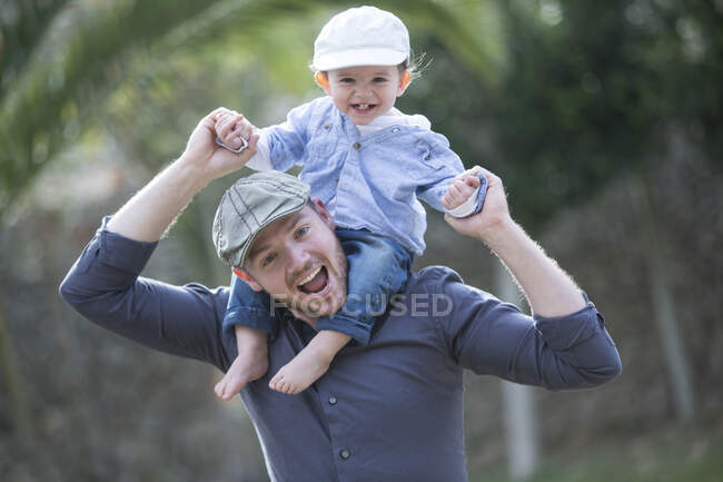 Bebé niño con gorra de béisbol sentado en los hombros del padre mirando a la cámara sonriendo - foto de stock