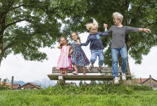 Nonna e nipoti a mezz'aria saltando dalla panchina del parco, Fuessen, Baviera, Germania — Foto stock