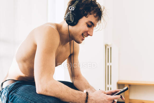 Nu peito homem vestindo fones de ouvido olhando para baixo no smartphone sorrindo — Fotografia de Stock