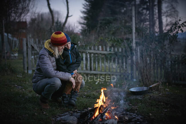 Mature woman and son crouching to watch garden campfire at dusk — Stock Photo
