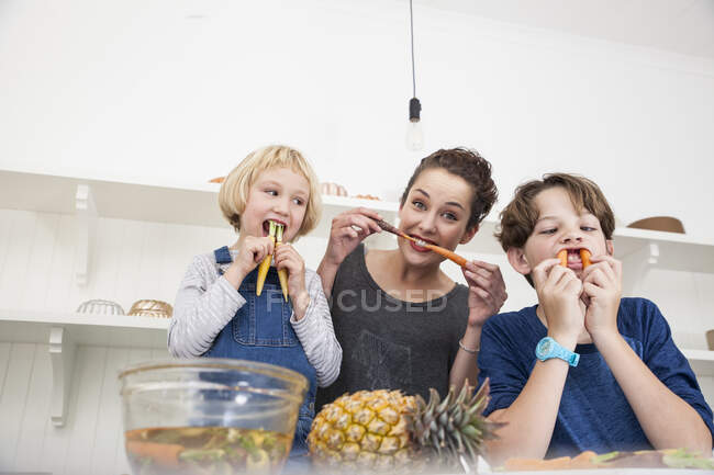 Young woman, boy and girl in kitchen, fooling around, using carrots as false teeth — Stock Photo