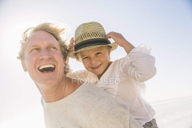 Pai dando filho piggyback sorrindo, usando chapéu de sol de palha — Fotografia de Stock