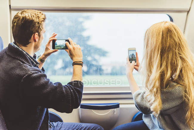 Pareja joven tomando fotografías de teléfonos inteligentes a través de la ventana del tren, Italia - foto de stock