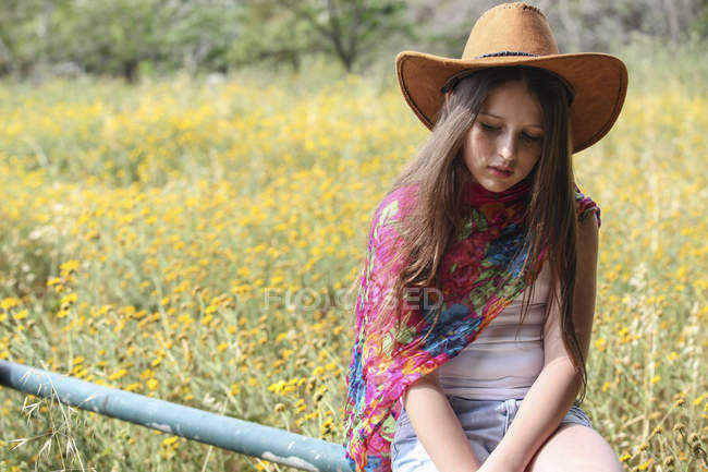 Sullen girl in cowboy hat sitting on fence gazing down — Stock Photo
