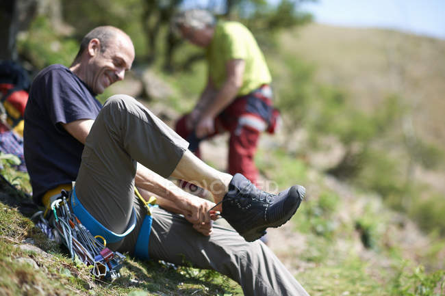 Hombre sentado en la ladera poniéndose una bota sonriente - foto de stock