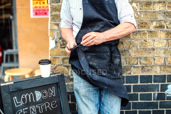 Senior craftsman outside workshop, cleaning glasses with his apron, mid section — Stock Photo