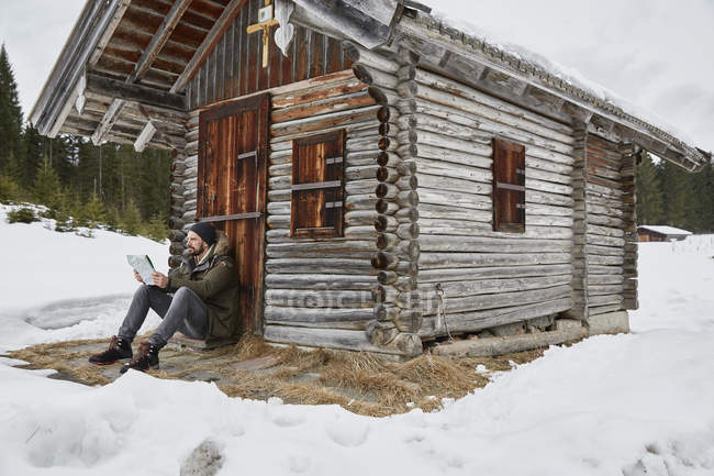 Young man reading map sitting outside log cabin in winter, Elmau, Bavaria, Germany — Stock Photo