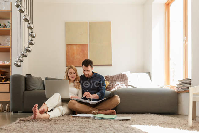 Пара с ноутбуком, глядя на документы, сидя на полу рядом с диваном — стоковое фото