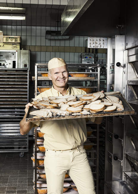 Happy baker placing tray of sliced bread into oven — Stock Photo
