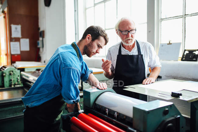 Artesano / técnico senior que supervisa al joven en la máquina de tipografía en el taller de artes del libro - foto de stock