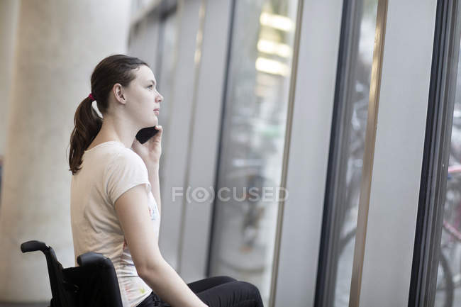 Young woman using wheelchair gazing through entrance window talking on smartphone — Stock Photo