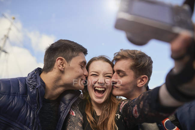 Tres amigos tomando selfie con cámara, hombres jóvenes besando a una mujer joven en la mejilla, Bristol, Reino Unido - foto de stock
