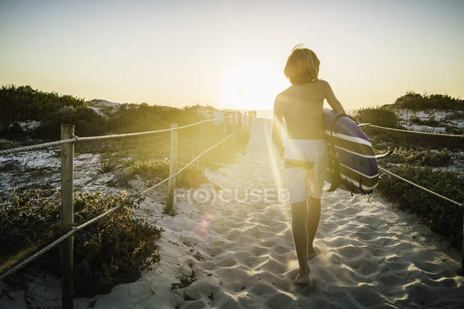 Young boy, walking along beach walkway, holding surfboard, rear view — Stock Photo