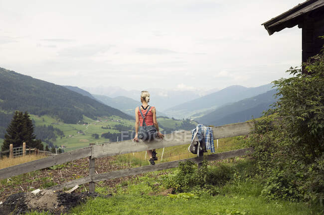Rückansicht einer am Zaun sitzenden Frau mit Blick auf die Berglandschaft, Sattelbergalm, Tirol, Österreich — Stockfoto