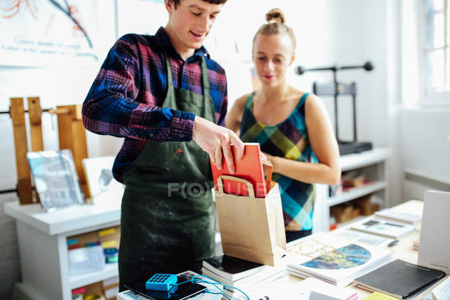 Young craftsman with customer, placing book into bag in arts bookshop — Stock Photo